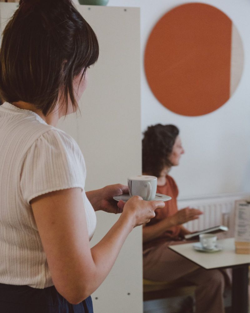 Frau mit weißem Oberteil trägt eine Tasse Kaffee zu einem Tisch im BuchQuartier-Café